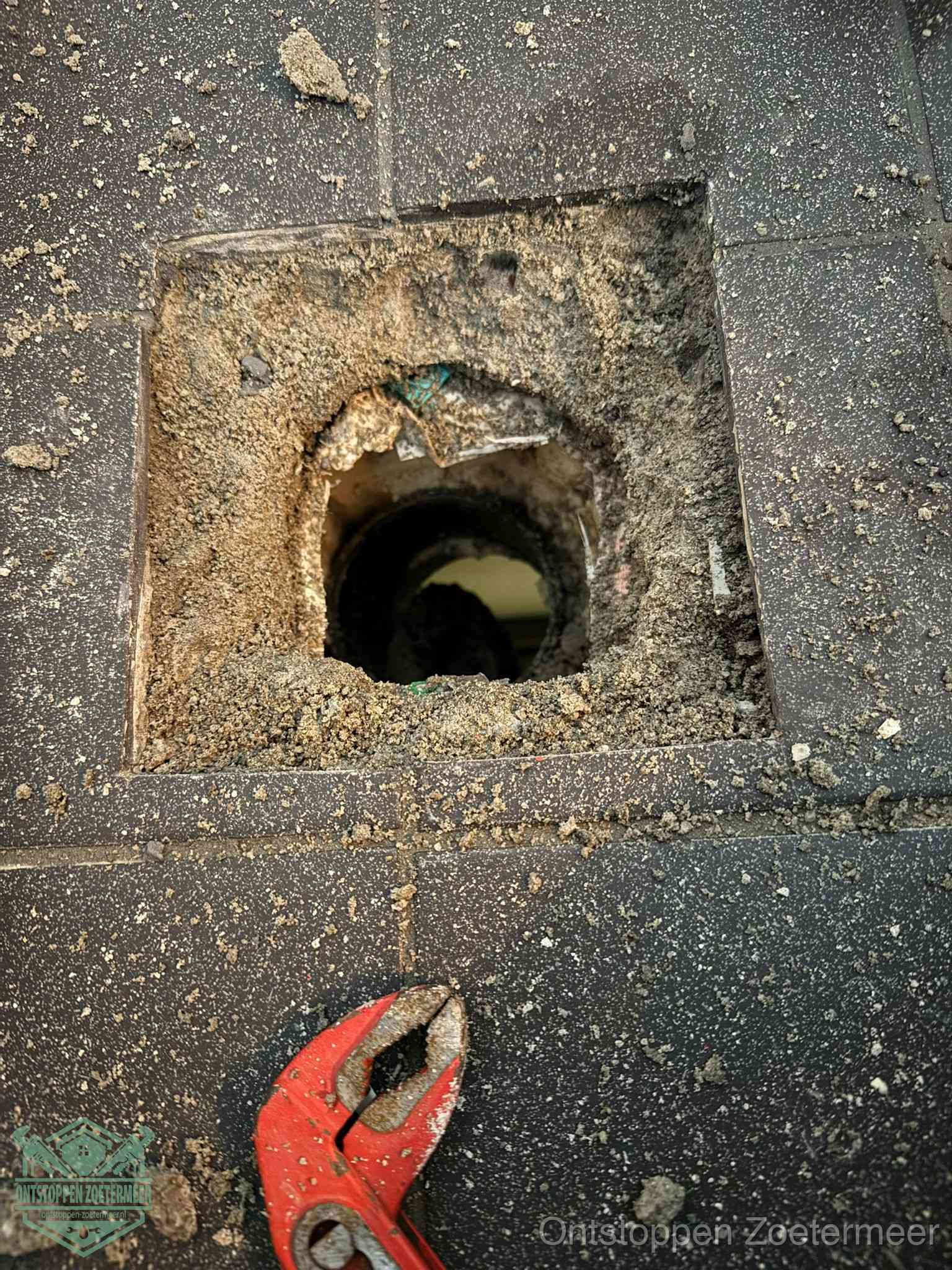 Een geopend rioolputje in een betontegelvloer met losgemaakt rooster en een zichtbare ronde afvoerbuis, het achterhalen waar de afvoerverstopping zit in Zoetermeer