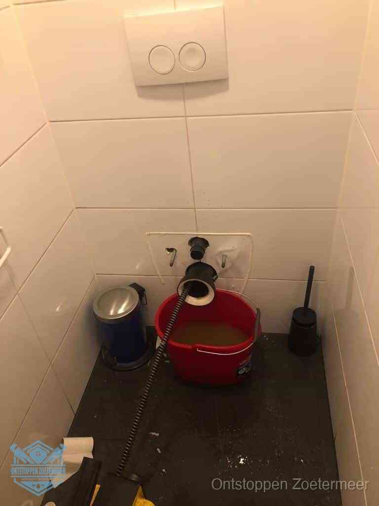 Een ontstopte wand-wc met verwijderde toiletpot waar een ontstoppingsveer in de wanduitgang verdwijnt, het achterhalen waar de verstopping zit in Zoetermeer