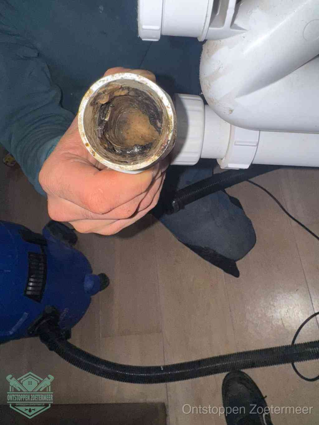 Een hand houdt een losgekoppeld PVC-afvoerbuisstuk vast dat van binnen volledig is dichtgeslibd met vet, vuil en aanslag, de meest voorkomende oorzaak van een verstopte afvoer in Zoetermeer