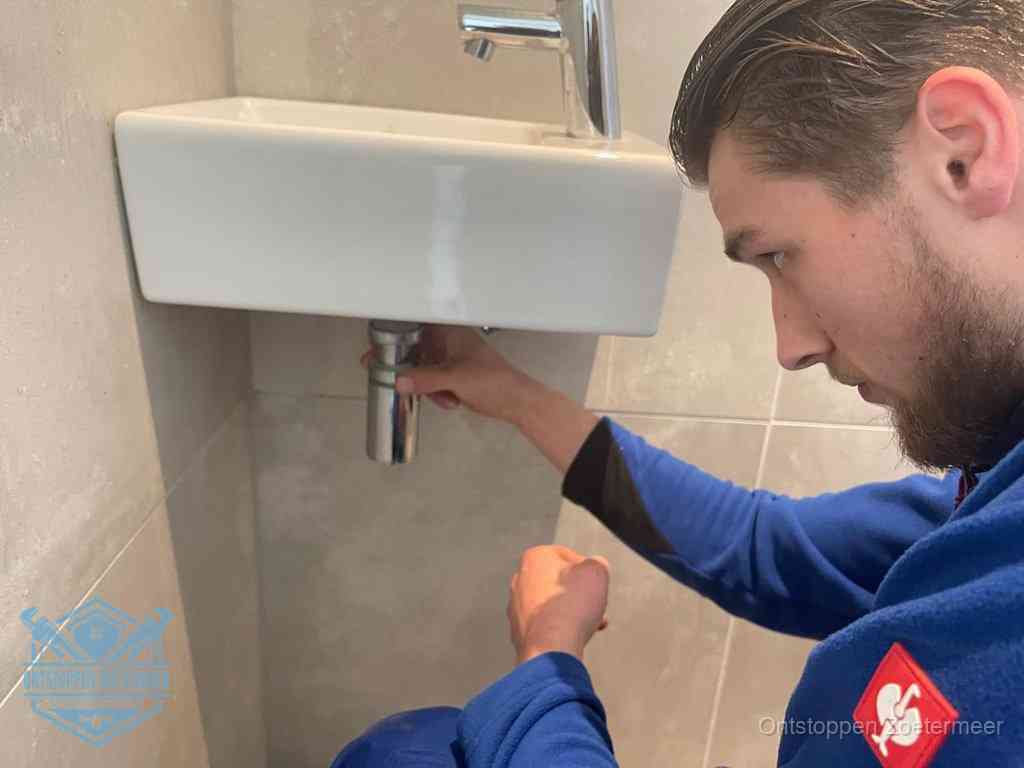 Een monteur draait met de hand de zwanenhalssifon onder een fonteinwasbak in de toiletruimte los om de bron van rioollucht in Zoetermeer te vinden