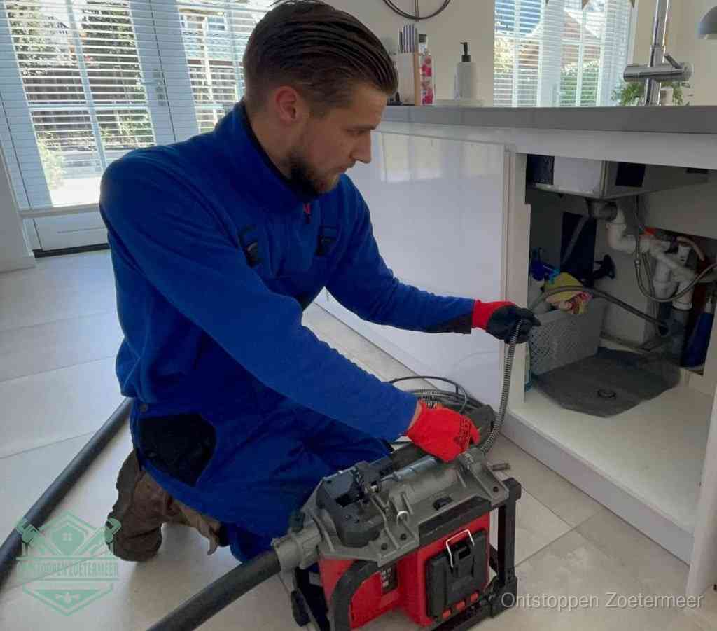 Een monteur met rode handschoenen knielt in een keuken en voert een elektrische ontstoppingsveer in tijdens het professioneel afvoer ontstoppen in Zoetermeer