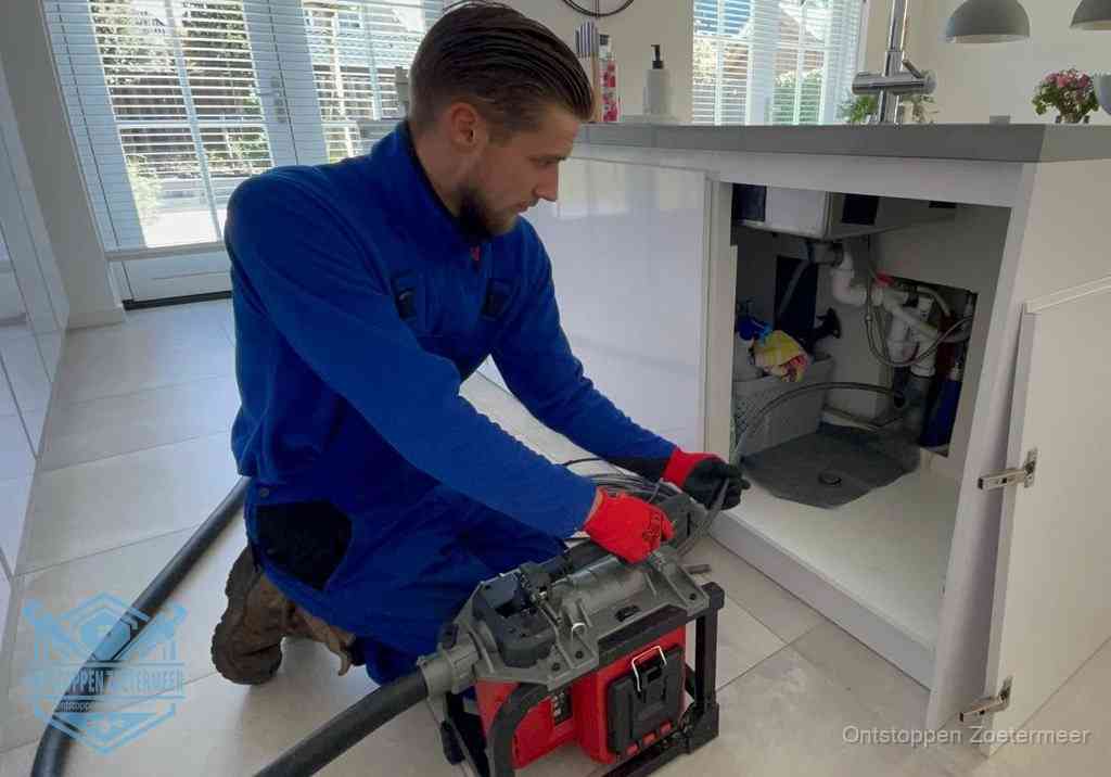 Een monteur voert een professionele elektrische ontstoppingsmachine in de afvoer onder de spoelbak tijdens het gootsteen ontstoppen in Zoetermeer